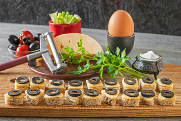 Sandwiches with cheese and black olives next to an egg, salt, tomatoes and herbs are laid out on a rustic kitchen table near a dark wall