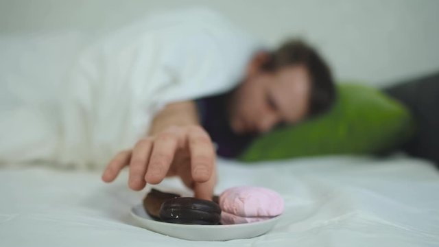 Poor diet concept. Man lies on the bed and reaches for a sweet cookie. Focus on plate with flour pastries. Unhealthy food. Caucasian white candyman covered with blanket eats lying down on green pillow