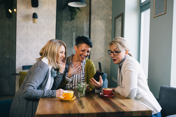 mid aged woman friends talking and drinking coffee in cafe, using mobile phone