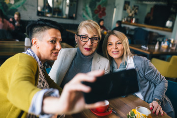 mid aged woman friends taking picture with mobile phone while drinking coffee in cafe