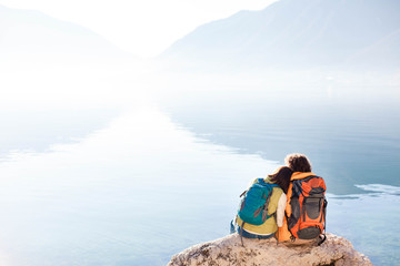 Couple travelers with hiking backpacks at sea beach. Tourists by blue lake and mountains. Young man and woman enjoying traveling, adventure, relaxation, silence and calmness. Copy space. Rear view.