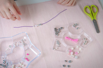 kid weaves a bead bracelet on a white table with a pink cloth and a container for beads in candy style