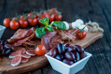 Prosciutto, bread, olives, walnut, mozzarella, salami, basil and cherry tomatoes on  brown wooden board.  Mediterranean kitchen.