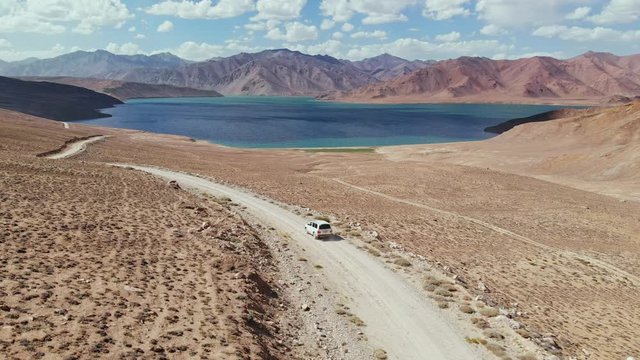 Aerial over off road 4x4 car driving along gravel trail path toward lake in arid mountains.Pamir Highway silk road travelling adventure in Tajikistan,Asia.Khorugh Bulunkul lake. 4k drone flight video