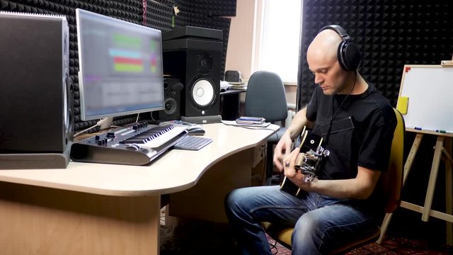 Bold male musician in black t-shirt and headphones plays guitar and records music with professional equipment in sound insulated recording studiod black 