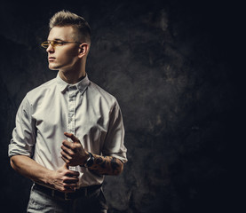 Fototapeta na wymiar Handsome and inked hipster male model posing in a studio wearing a white shirt, yellow glasses, standing in front of the grey background, looking cool and interested