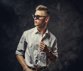 Interested and inked hipster male model posing in a studio, wearing sunglasses, white shirt, standing in front of the grey background, looking cool and confident
