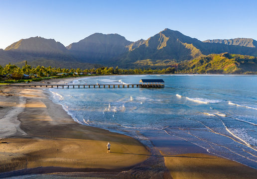 Aerial image at sunrise off the coast over Hanalei Bay and pier on Hawaiian island of Kauai with a man standing alone on the beach
