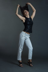 a brunette in a black leather sleeveless blouse and white trousers poses on a gray background