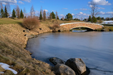 Fototapeta na wymiar The landscape is early spring. The landscape of the city park with melting snow on hills, trees, shrubs, a bridge over the lake covered with ice and a blue sky with clouds.