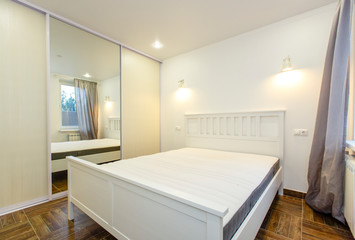 bedroom in white colors in the house. Large white double bed, large mirror on the sliding wardrobe, beige parquet floor