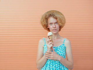 young cheerful curly redhead woman in straw hat and blue sundress eating ice cream on beige background. Fun, summer, fashion, youth concept