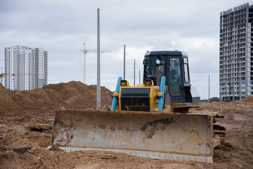 Dozer during land clearing and foundation digging against tower cranes at large construction site. Bulldozer with bucket for pool excavation and utility trenching. Earthmoving works & heavy machinery