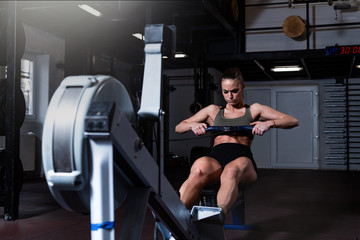 Young strong fit sweaty powerful attractive muscular woman with big muscles doing hard core row heavy cross training workout on indoor rower at the gym real people exercise