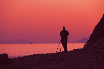 Fototapeta na wymiar Silhouette of a male photographer by the sea during sunset it makes picturesque pictures of hills and landscapes on sunny summer evening. Nature photography concept. Advertising space
