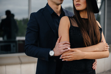 A beautiful, stylish pair of young people in black clothes and glasses stand against the background of an office building in the sunset. Fashion and style