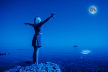 Fototapeta na wymiar Rear view unidentified young happy woman stands on a stone raise Hands up Looking at the big moon and calm clear sea water against the backdrop of landscape and clear night sky. Full moon concept