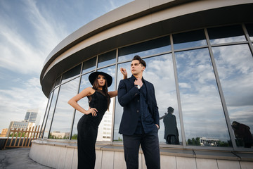 A beautiful, stylish pair of young people in black clothes and glasses stand against the background of an office building in the sunset. Fashion and style