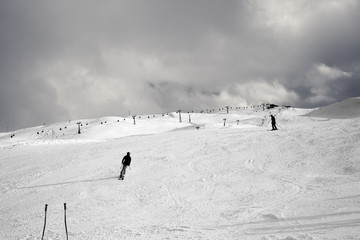 Snowy ski slope with skiers in high mountains and cloudy sunlit sky at winter day