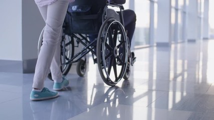 Low-section shotof nurse pushing driving wheelchair with handicapped patient through the corridor with mirror floor - Powered by Adobe