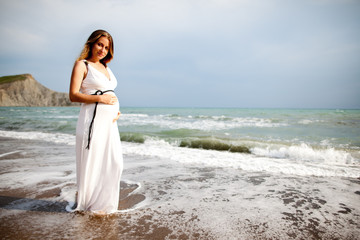 Pregnant lady with brown hair dressed in white dress, standing on beach near water. On background mountain and sea