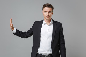 Serious young business man in classic black suit shirt posing isolated on grey wall background. Achievement career wealth business concept. Mock up copy space. Showing stop gesture with palm aside.