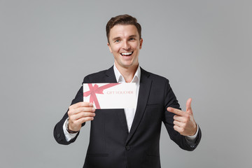 Funny young business man in classic black suit shirt posing isolated on grey wall background. Achievement career wealth business concept. Mock up copy space. Pointing index finger on gift certificate.