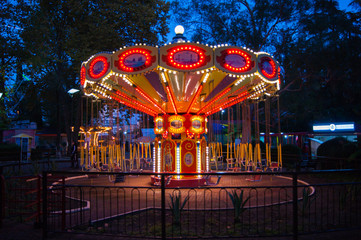 Carousel in bright lights on a dark summer south night against a background of trees and other attractions.