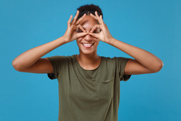 Naklejka premium Funny young african american woman girl in casual t-shirt posing isolated on blue background. People lifestyle concept. Mock up copy space. Holding hands near eyes imitating eyeglasses or binoculars.