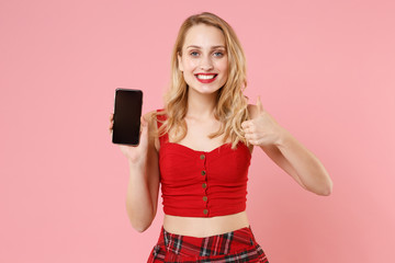 Naklejka premium Smiling young blonde woman girl in red sexy clothes posing isolated on pastel pink background. People lifestyle concept. Mock up copy space. Hold mobile phone with blank empty screen showing thumb up.
