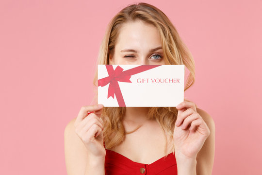 Funny young blonde woman girl in red sexy clothes isolated on pastel pink wall background studio portrait. People lifestyle concept. Mock up copy space. Covering face with gift certificate blinking.