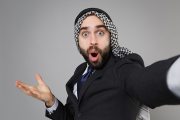 Fototapeta premium Close up of arabian muslim businessman in keffiyeh kafiya ring igal agal suit isolated on gray background. Achievement career wealth business. Doing selfie shot on mobile phone keeping mouth open.