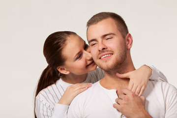 Front view of charming young woman hugging joyful guy from behind and biting ear. Handsome man holding girlfriend hand and smiling. Isolated on white studio background. Concept of relationships.