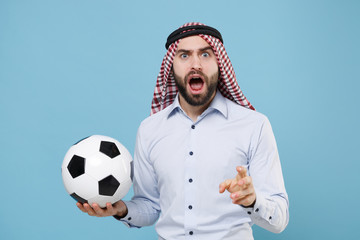 Perplexed arabian muslim man in keffiyeh kafiya ring igal agal casual clothes isolated on pastel blue background. People religious lifestyle concept. Cheer up support favorite team with soccer ball.