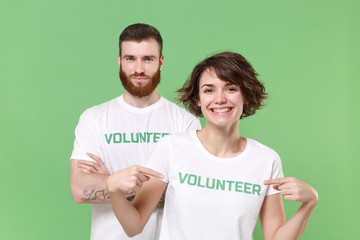 Smiling two friends couple in white volunteer t-shirt isolated on pastel green wall background. Voluntary free work assistance help charity grace teamwork concept. Point index fingers on themselves.