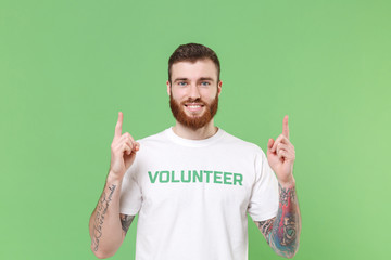 Smiling young bearded man in white volunteer t-shirt isolated on pastel green background studio...