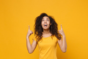 Excited young african american woman girl in casual t-shirt posing isolated on yellow orange wall background studio portrait. People lifestyle concept. Mock up copy space. Pointing index fingers up.