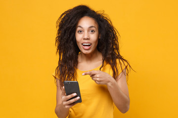 Amazed young african american woman girl in casual t-shirt isolated on yellow orange background. People lifestyle concept. Mock up copy space. Point index finger on mobile phone, typing sms message.