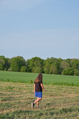 Boy with long hair, black t-shirt and blue shorts walking on a green field with trees in the background.
