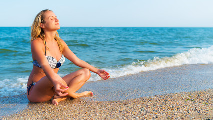 Fototapeta na wymiar Woman in white and blue swimsuit sits on seashore in yoga pose with her eyes closed. Blurred background