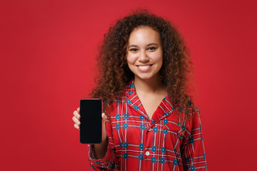 Smiling young african american girl in pajamas homewear resting at home isolated on red background. Relax good mood lifestyle concept. Mock up copy space. Hold mobile phone with blank empty screen.