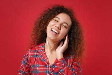 Pretty african american girl in pajamas homewear resting at home isolated on red background. Relax good mood lifestyle concept. Mock up copy space. Listen music with earphones keeping eyes closed.