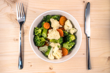 White vegetable bowl with carrot cauliflower and broccoli on a wooden base and cutlery.. Healthy, selective Focus