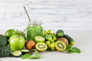 Various green fruits and vegetables with mason jar of smoothie on white wooden table over white brick wall. Detox diet.