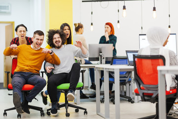 Fototapeta premium Colleagues having fun on work at the office. Girls pushing boys on chairs with wheels.