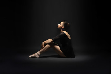 Beautiful young girl ballerina in a black dress and pointe shoes sits on a black background in the rays of light