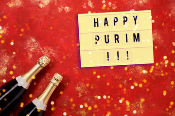 Happy Purim written in light box and champagne bottles on bright red background. Purim background. Copy space, top view