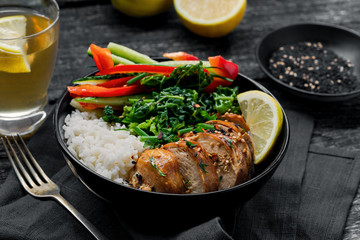 Buddha bowl meal with teriyaki chicken fillet, fresh vegetable salad, stew cabbage and basmati rice on a rustic table. Delicious dish in a black bowl with homemade lemonade.