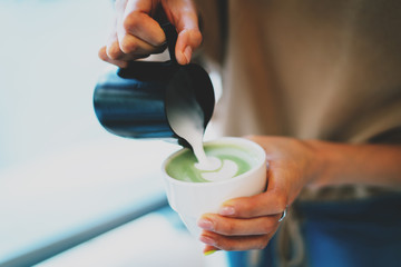 Closeup photo of a woman's hands pouring a milk into a freshly made matcha tea. Preparing japanese green tea with a milk. Female barista making matcha latte. Matcha green tea with a hot milk.
