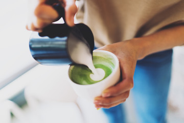 Professional female barista adding whipped milk to a freshly made matcha tea. Woman's hands holding a cup of a matcha tea and a jug with a hot whipped milk. Adding whipped milk to the japanese tea.